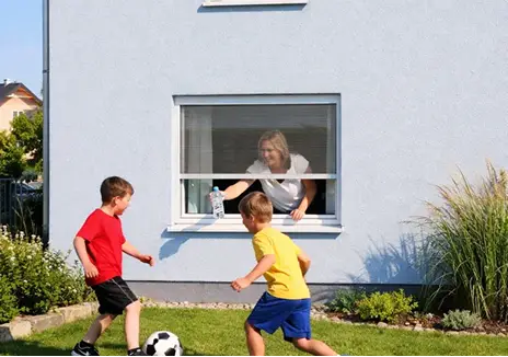 Fliegengitter-Rollo am Fenster für zuverlässigen Insektenschutz Frau öffnet ein Fliegengitter-Rollo am Fenster, während zwei Kinder im Garten Fußball spielen