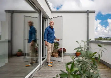 Fliegengitter-Drehtür nach Maß für Balkon und Terrasse Mann öffnet eine maßgefertigte Fliegengitter-Drehtür auf einer Terrasse mit Pflanzen und Glasfront